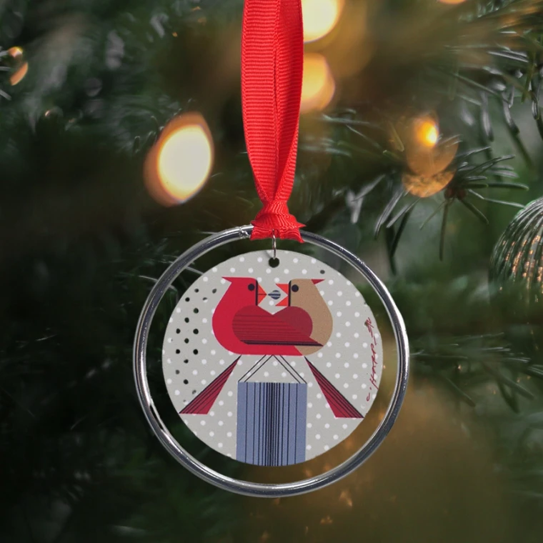This circular double-sided metal ornament features the 1970 "Redbirds Romance" illustration of a  male cardinal feeding a female cardinal a berry in front of a polka-dotted background, in the "minimal realism" and geometric style Charley Harper was known for. A silver ring hangs separately, with a red ribbon attached to this for hanging.