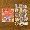 Cat's Atelier package with individual die-cut cat and artist studio stickers on a wood grain background