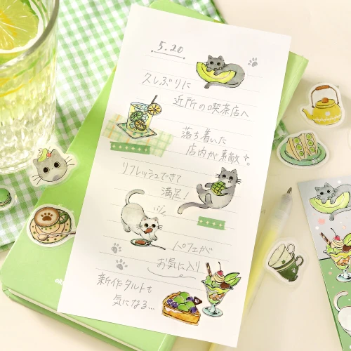 Gray Cat Welcome Meow stickers decorating a journal page on a green gingham surface
