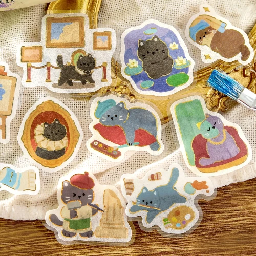 Cat's Atelier stickers of framed cat portraits and cat artists arranged on a woven fabric surface