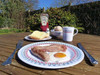 Image of a breakfast served on Large Witley Melamine plate, mug of tea and small plate with a buttered roll on summers day on an outdoor table.