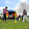 A pair of young woman play swingball in front of the orange Uno  Breeze® v2 Campervan Awning whilst their friends sit outside the awning in camping chairs ,imageset:Orange
