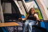 A woman sits inside the the OLPRO Cocoon V2 Poled Campervan Awning by OLPRO she is sat in an OLPRO camp chair next to a lightweight bamboo table