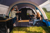 A group of friends sit inside the Poled Cocoon V2 Campervan Awning by OLPRO, some sat in camping chairs surrounding a bamboo folding table