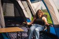 A woman sits inside the the OLPRO Cocoon V2 Poled Campervan Awning by OLPRO she is sat in an OLPRO camp chair next to a lightweight bamboo table