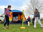 A pair of young woman play swingball in front of the orange Uno  Breeze® v2 Campervan Awning whilst their friends sit outside the awning in camping chairs ,imageset:Orange