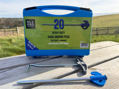 A case of 20 OLPRO Hard Ground Pegs, featuring a single peg and the accompanying peg puller, displayed on a table against the backdrop of the expansive countryside.
