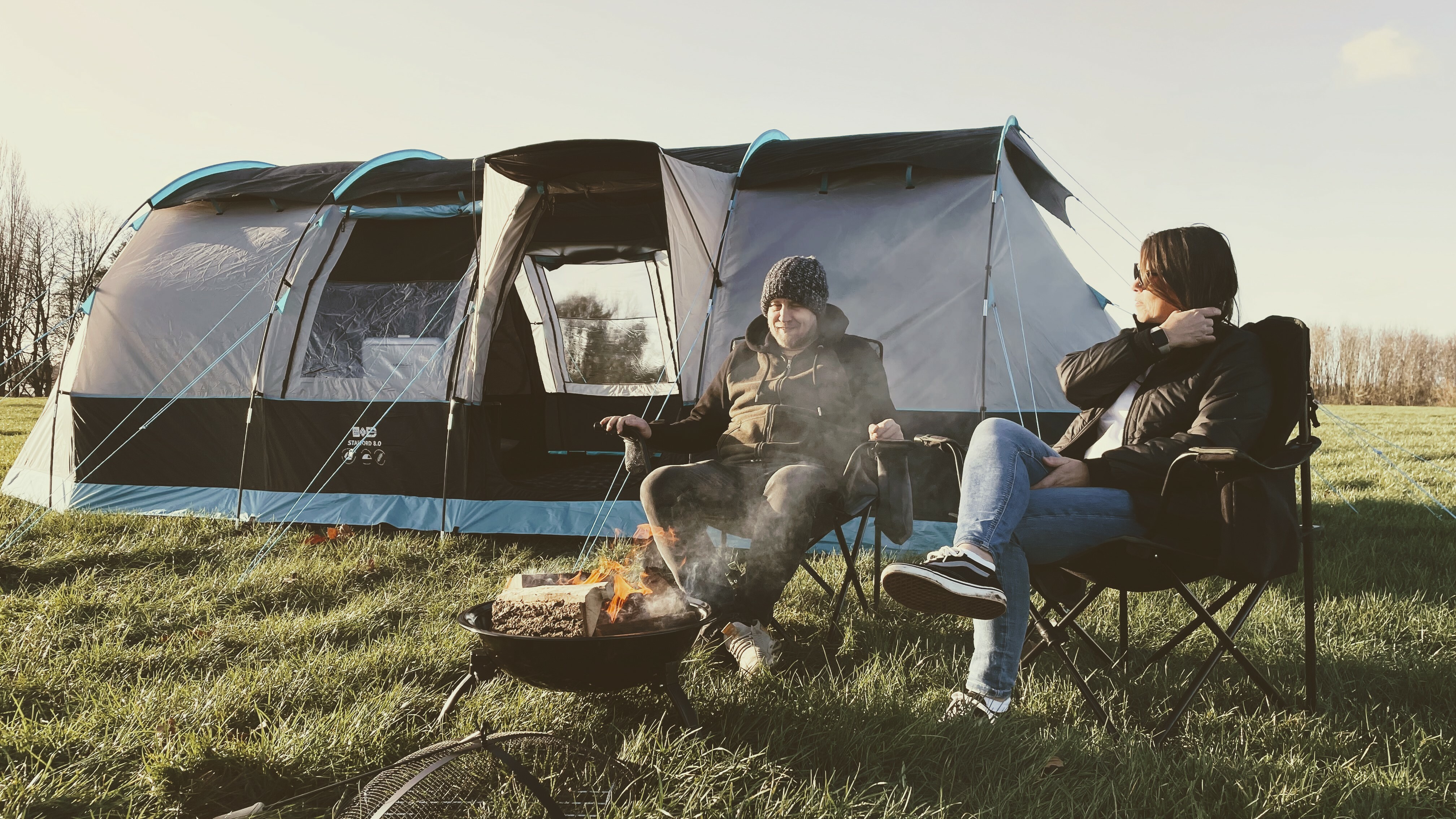 A couple sit by a fire in camping chairs, behind them is the Stafford 8.0 Family Tent