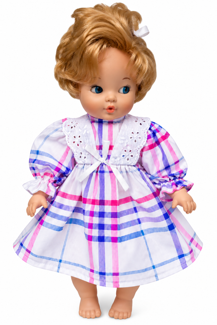 Front view Adorable blonde baby doll wearing a pink, purple, and blue plaid dress with puff sleeves, white eyelet lace collar, ruffled cuffs, and a white satin bow. Doll stands in a charming pastel nursery with flowers, a pink teddy bear, and a gift box. Handmade-style dress for little baby dolls from Doll Clothes Superstore.