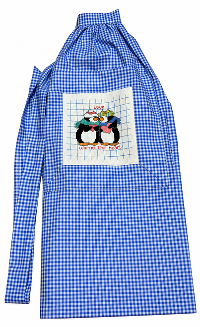 Handmade blue and white gingham half apron with gathered waist, long waist ties, and front pocket featuring two embroidered penguins with the words “Love Warms the Heart.” Shown worn over a white shirt and jeans. Available from Doll Clothes Superstore. Perfect hostess gift