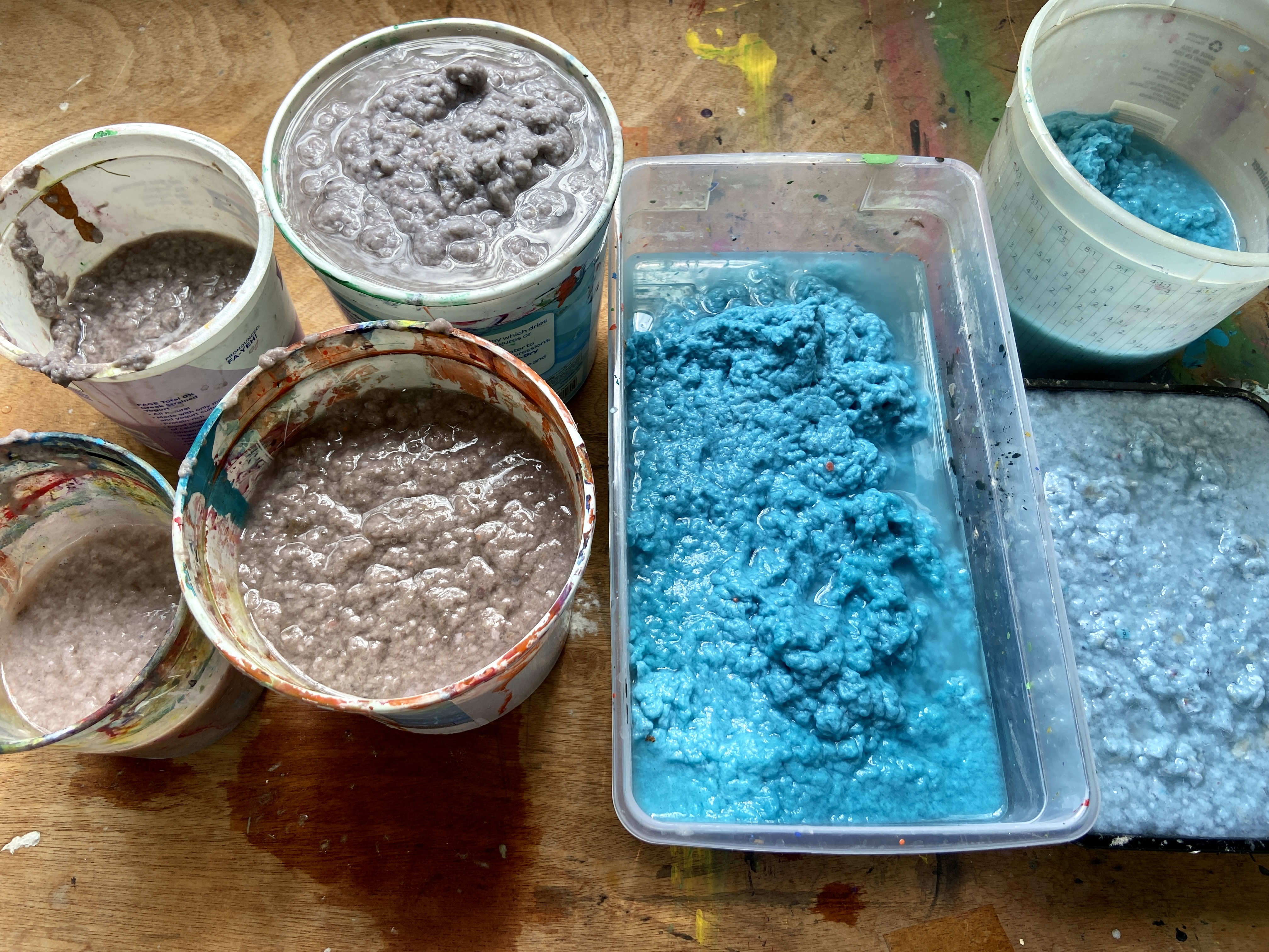 Paper pulp in seven containers, colors ranging from multiple shades of blue and tans