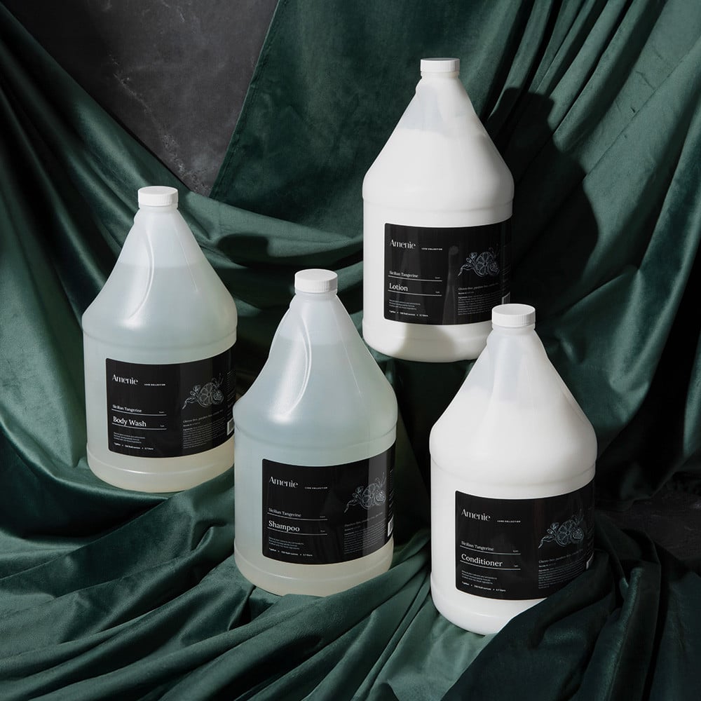 Amenie Sicilian Tangerine complete gallon collection with shampoo, conditioner, body wash, and lotion on green tropical leaf backdrop