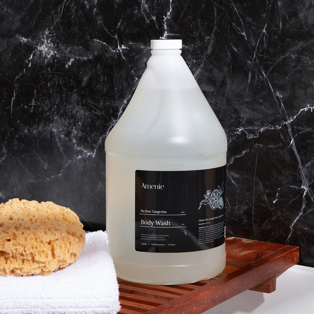 Amenie Sicilian Tangerine Body Wash gallon-size bottle with sponge and orange slices on black marble backdrop