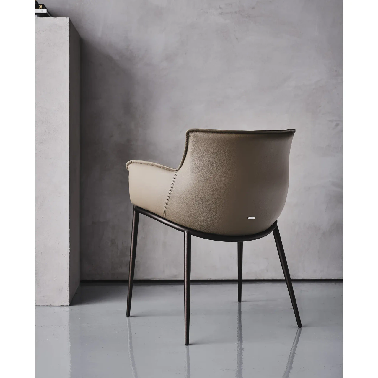 FANGO || BRONZE EMBOSSED STEEL - Modern taupe leather armchair with slim black metal legs, rear three-quarter view against a grey concrete wall.