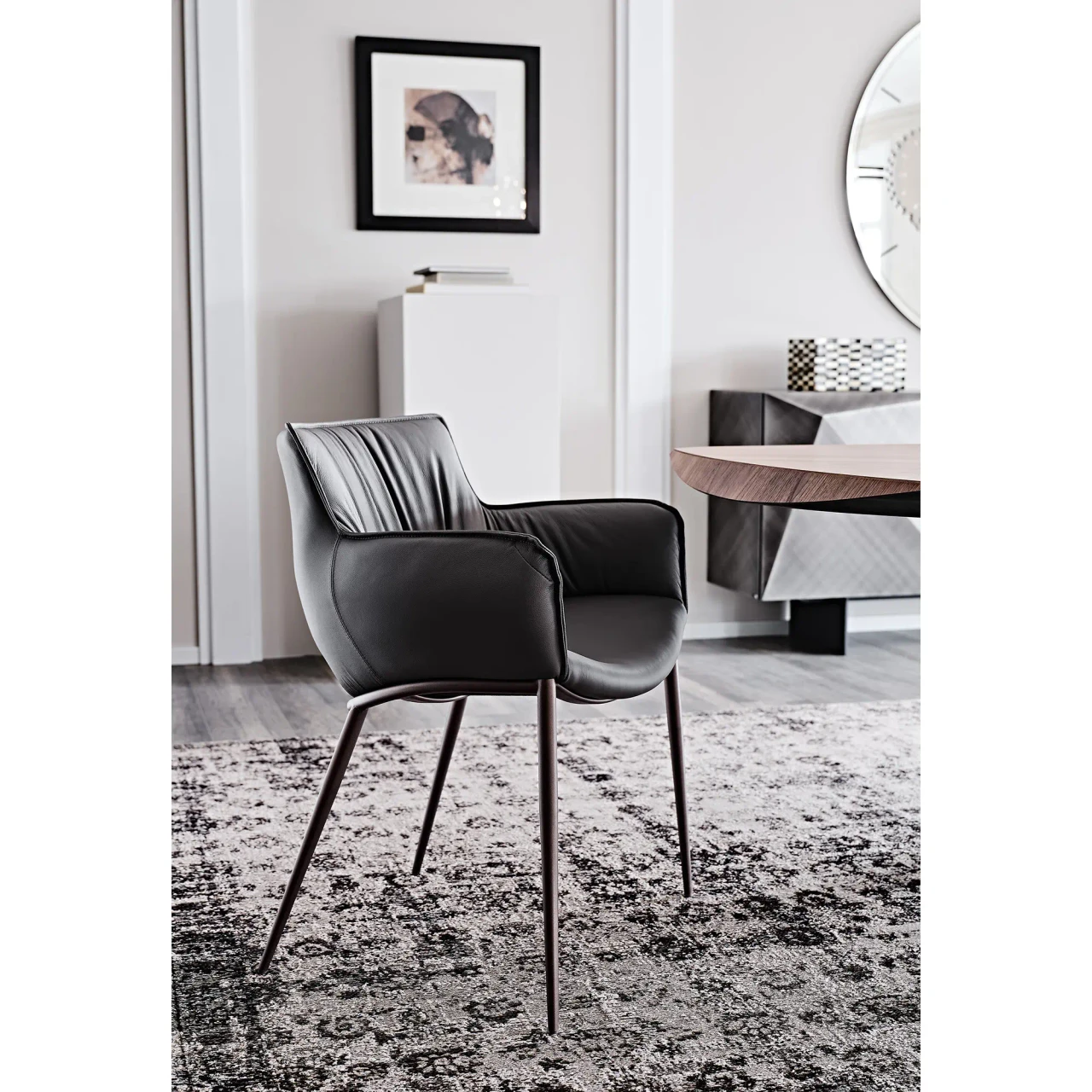 SMOKE || GRAPHITE EMBOSSED STEEL - Modern black leather armchair with tapered dark wood legs, side view, set in a contemporary living room.