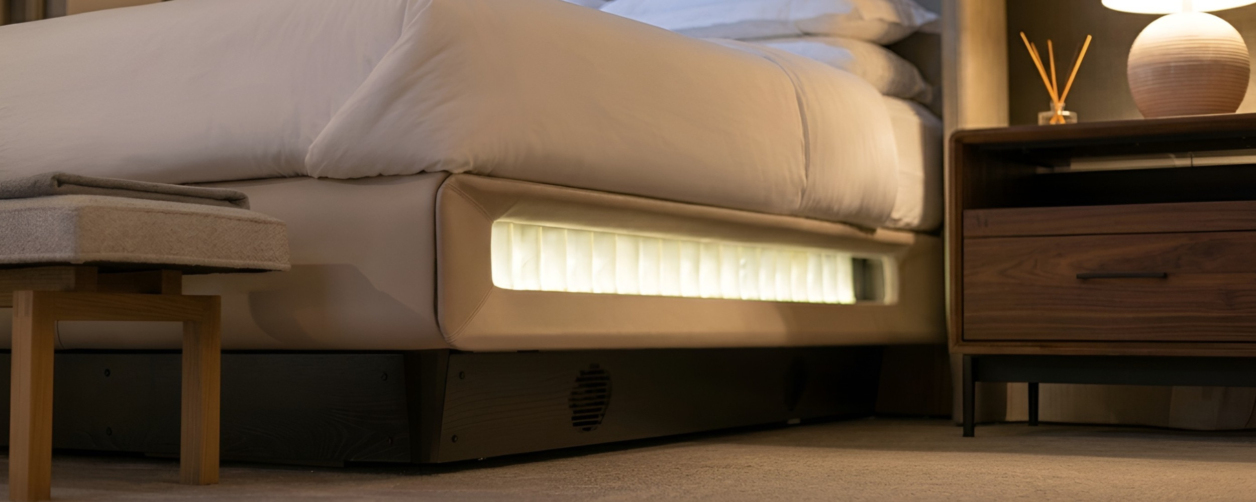 An angled low-view photograph, from floor level, shows a made bed with white bedding next to a two-drawer wooden nightstand, against a grey-toned textured wall.