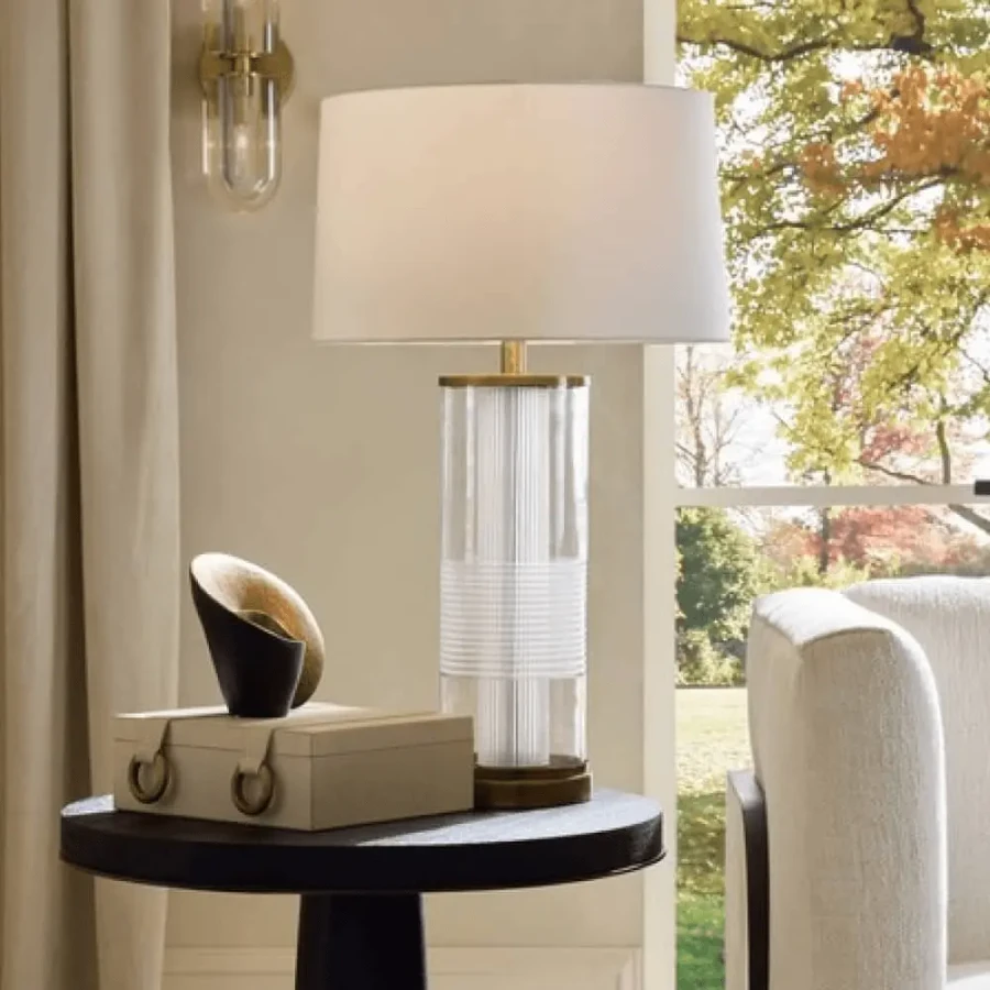 Gianna Table Lamp with brass accent, clear etch glass base, and off-white cotton shade in a living room, eye-level view.