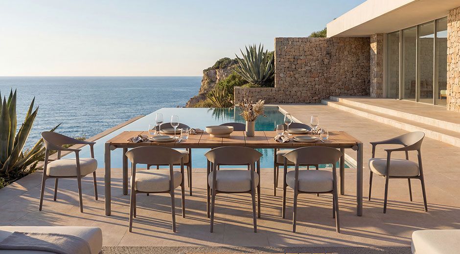Luxury outdoor dining set with a wooden table and grey upholstered chairs on a patio next to an infinity pool overlooking the ocean.