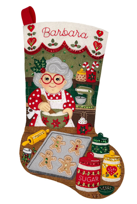 Personalized felt Christmas stocking kit featuring Mrs. Claus baking holiday treats in a festive kitchen. The design includes gingerbread cookies, candy canes, baking ingredients, kitchen accessories, and colorful holiday decorations, with a customizable name banner across the top. Handcrafted felt details are accented with sequins and beads in traditional red, green, and white Christmas colors