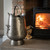 Antique pewter coal bucket on a stone hearth beside a lit wood-burning stove with a stacked log pile