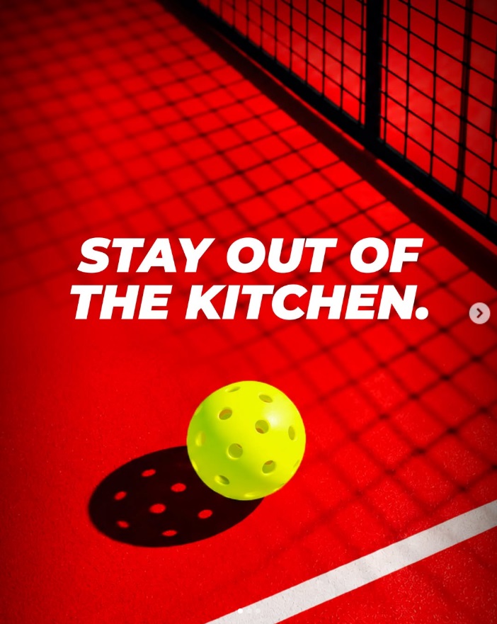 Yellow pickleball on a red court beside a net with the words Stay out of the kitchen