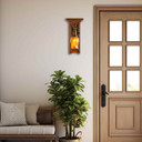  A tall, wall mounted sconce lantern made with quarter sawn oak like Stickley furniture. The fixture is mounted on a metal hook with an aged brass finish, turn key socket and a curvy, blown glass cherry blossom shade. It is installed above a houseplant next to a front door.