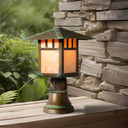  A linear, formal Craftsman lantern with an aging patina finish and warm glass on a brass post sits on a porch railing inviting guests. This fixture has a window pattern artwork reminiscent of the Arts & Crafts era.