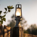  An antique-style post-mount lantern with a glowing frosted glass shade, installed on top of a weathered wooden fence post at dusk.