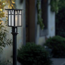  A rectangular, vertical Craftsman style post mounted light fixture with a black finish and warm white glass in front of an out of focus house with climbing foliage.
