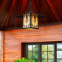  A linear Arts & Crafts chain mounted pendant light fixture with a black finish, warm colored glass and the silhouette of a giant Sequoia tree. The lantern is hanging from the ceiling of a home exterior near a window.