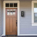  A solid brass, vertically oriented illuminated house number plaque with a two number address, a satin black finish, warm-toned glass and corner glass accents installed on the siding of a house next to the front door.