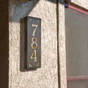  A solid brass, vertically oriented illuminated house number plaque with a three number address, a bronze finish, warm glass and corner glass accents installed on a stucco wall near a pair of windows.