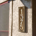  A solid brass, vertically oriented illuminated house number plaque with a four number address, an aged finish, iridescent glass and corner glass accents installed on a stucco wall near a pair of windows.