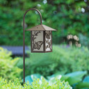  An artistic garden lantern on a shepherd's hook post with smooth bronze finish, butterfly artwork and white glass installed in shrubs in a backyard.