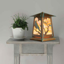  A beefy, rustic pedestal style table lamp with Gothic literature raven silhouettes in trees, an aged patina finish and iridescent glass. It sits on a gray nightstand next to a bed with white pillows and comforter.