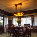  An elegant chandelier inspired by the work of Charles and Henry Greene in the Gamble House in Pasadena, California. It has a linear design, leather strap suspension, wood body with ebony accents and an iridescent art glass vine design. It's installed in a California Craftsman style dining room with neutral tones.