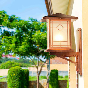  A Prairie School style perch sconce with a raw copper finish and white glass mounted to the side of a garage. There is a geometric, diamond accent pattern over the glass panels. Trees and shrubs from the landscaping around the garage are out of focus in the background.