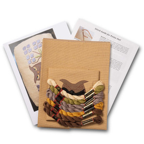  Overhead view of a embroidery kit for a petite owl pillow design, showing folded natural linen stamped with a owl pattern alongside arranged embroidery thread skeins in a variety of warm colors.