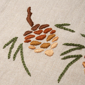  Closeup photo of a linen beige table scarf featuring embroidered artwork of a pine cone design.