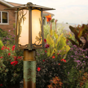  A rustic, artistic post mounted landscaping light fixture with a peaked roof, dragonfly artwork, an aged patina finish and warm glass installed in the brush and shrubs of a home property.