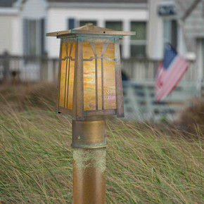  A linear Arts & Crafts post mounted light fixture with an aged patina, golden glass and a Gamble House design in front of waving grass and houses.