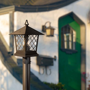  A traditional lantern with candle light bulbs and latticework windows sits on a post in front of a storybook cottage home.