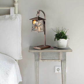  A beefy, rustic table lamp on a hooked arm with Halloween or horror film artwork of flying bats, a raw copper finish and custom black glass. It sits on a gray nightstand next to a bed with white linens.