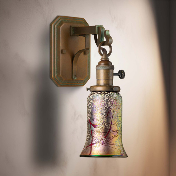  A reproduction of an antique Craftsman wall sconce light fixture with a metal corbel and hook mount for the lantern, which has a turn key socket, aged patina finish and a trumpet shaped cherry blossom blown glass shade. It's installed on an interior wall.