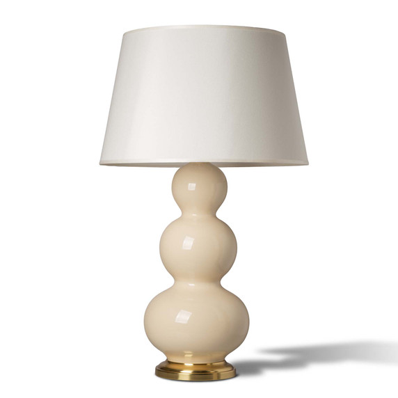  A bone colored Mid-Century table lamp featuring a glossy base with three stacked bulbous sections. The lamp is mounted on a polished brass round pedestal and topped with a slightly tapered off-white fabric drum shade.