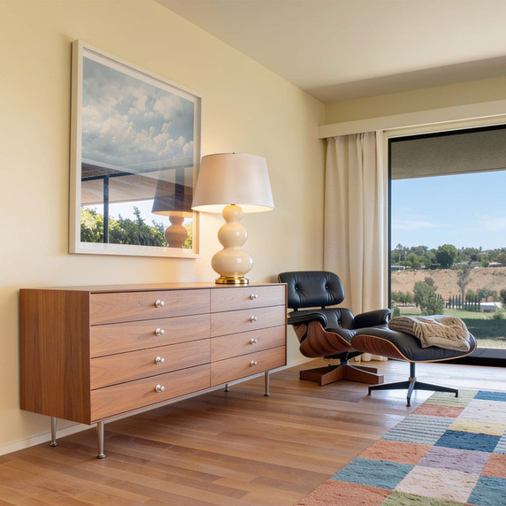  A vintage Triple Gourd cream ceramic table lamp with a white shade sitting on a mid-century modern wooden dresser next to an Eames lounge chair and ottoman.
