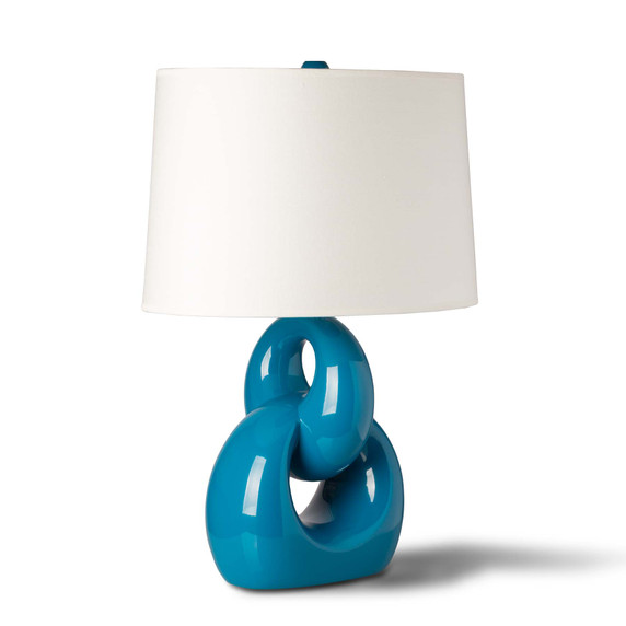   A mid-century modern style table lamp featuring a glossy peacock blue ceramic base designed as two interlocking rings. The sculptural base is topped with a simple white fabric drum shade against a plain white background.