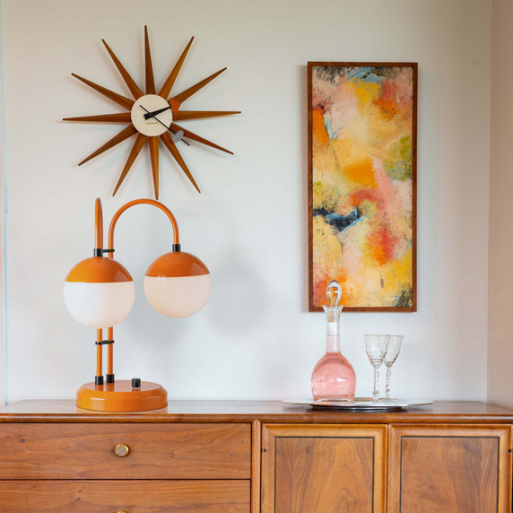  A mid-century lamp with an orange finish and two white globes sits on a credenza with modern art and accents.