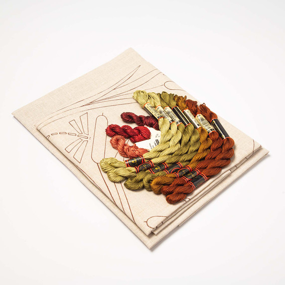  DIY embroidery kit for a dragonfly pillow design, showing folded natural linen stamped with a dragonfly and cattail pattern alongside arranged embroidery thread skeins in red, rust, brown, and olive green.