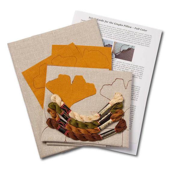  Overhead view of a embroidery kit for a gingko pillow design, showing folded natural linen stamped with a gingko leaf pattern alongside arranged embroidery thread skeins in beige, olive green, rust and brown with instructions.